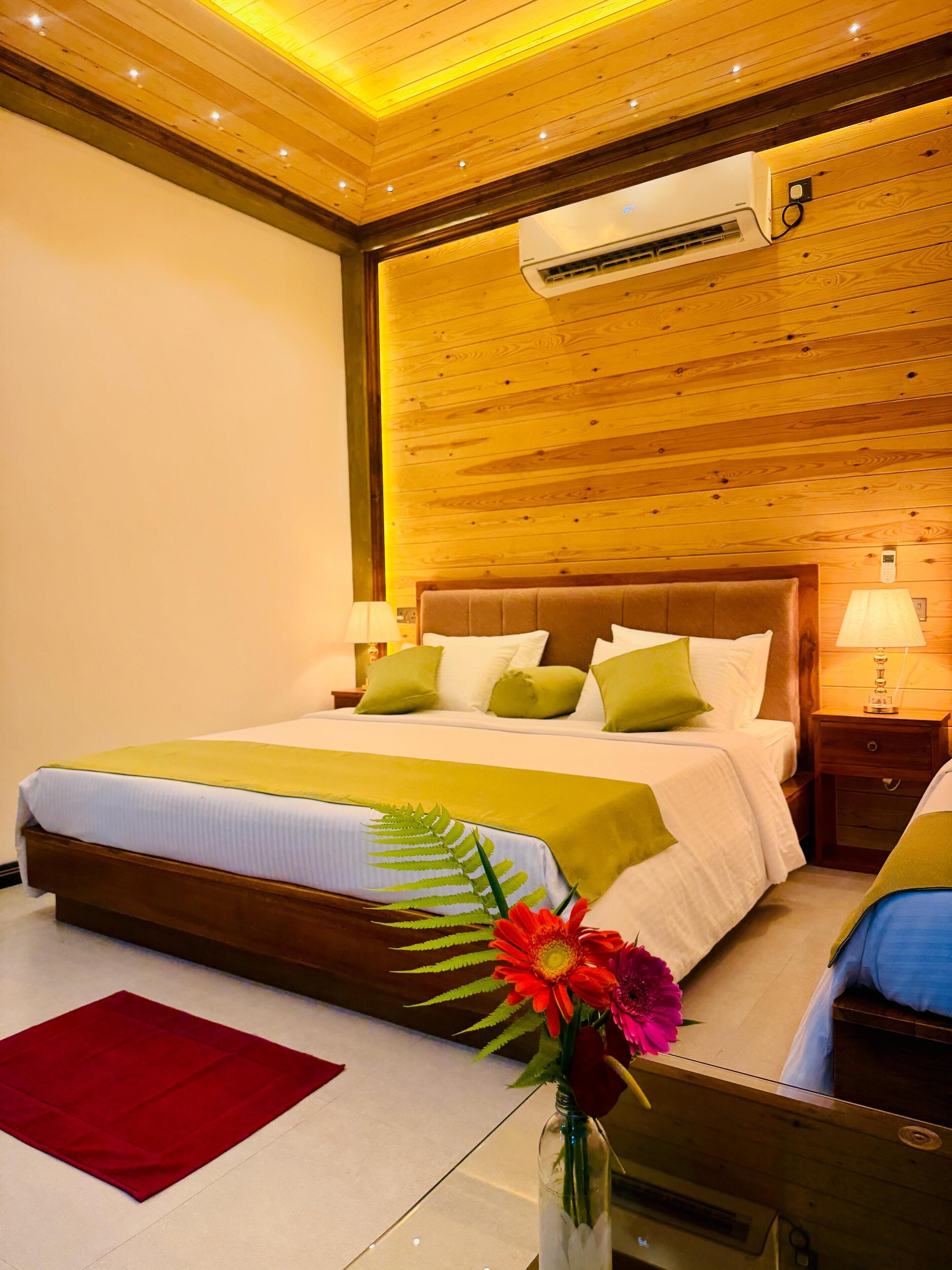 Habitación de hotel con techo de madera, una cama grande con ropa de cama verde y blanca, una mesita de noche con una lámpara y un jarrón con flores en el suelo.