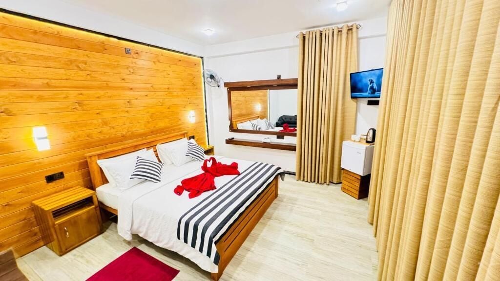 Habitación de hotel con cabecera de madera, cama con ropa de cama a rayas, una toalla roja, un televisor en la pared y un espejo.