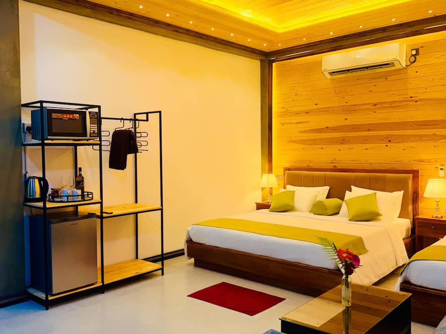 A modern hotel room with a wooden ceiling, a bed with multiple pillows, a small table with a vase, and a built-in air conditioning unit.