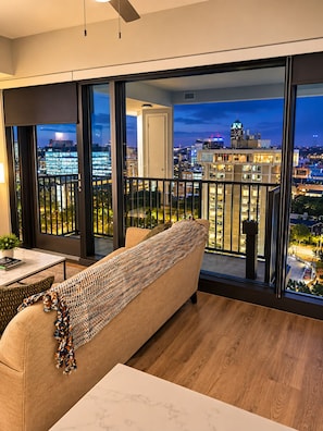 A modern living room with a sofa and a table, large windows with a city view, and a balcony with a railing. - Downtown Raleigh - Sweeping City Views - Luxury High Floor Unit 2 Bedroom 2 bath (Raleigh)
