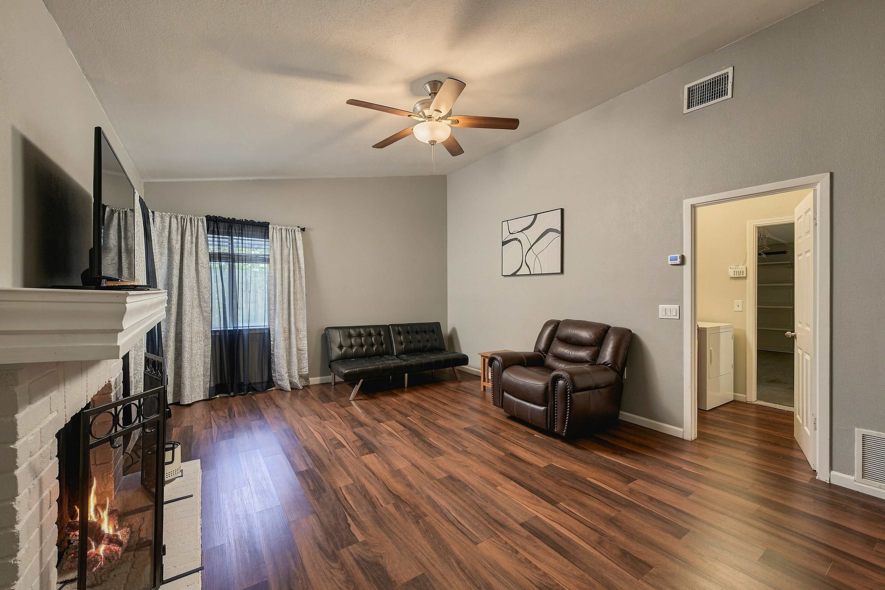 A living room with hardwood flooring, a fireplace, a ceiling fan, a black leather sofa, a leather armchair, and a painting on the wall.