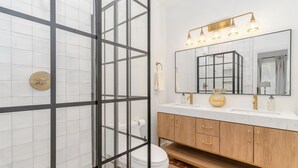 Ein Badezimmer mit einer Duschkabine aus Glas, einem Holzbadezimmermöbel mit goldenen Griffen und einem großen Spiegel mit einer Leuchte darüber.