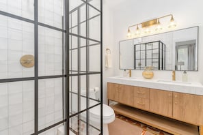 A bathroom with a glass shower enclosure, a wooden vanity with gold handles, and a large mirror with a light fixture above. - The Cowtown Cottage - 3-bedroom/3-bath house near Magnolia in Fort Worth (Fort Worth)