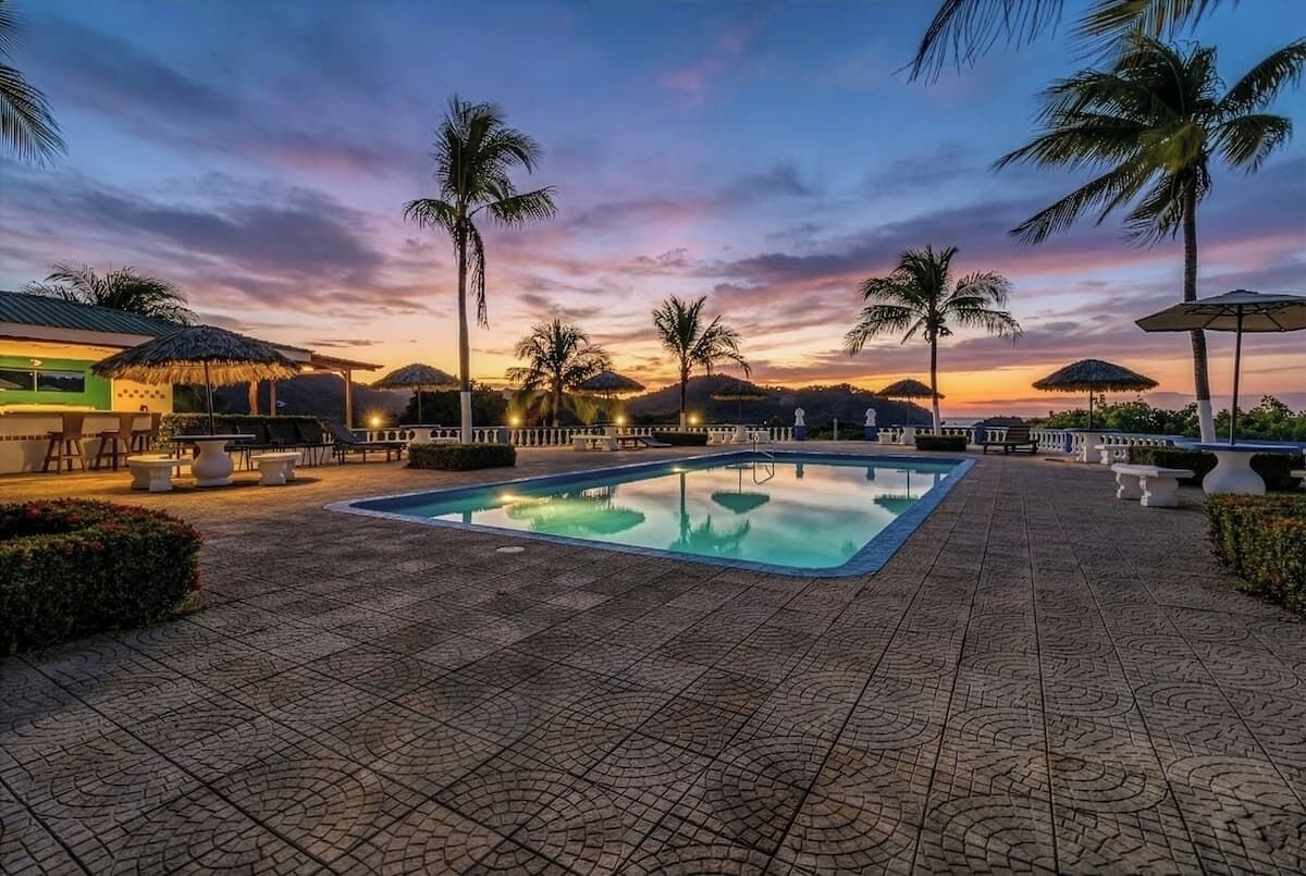 A swimming pool with a tiled deck, surrounded by palm trees and umbrellas, under a sunset sky.