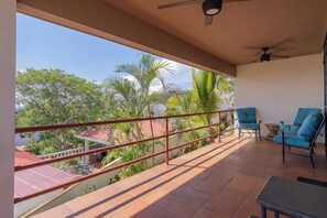 A balcony with a view of trees and a building, a ceiling fan, and blue chairs. - Resort Pool and Sunset Views: 2BR in Playa del Coco (Playa del Coco)