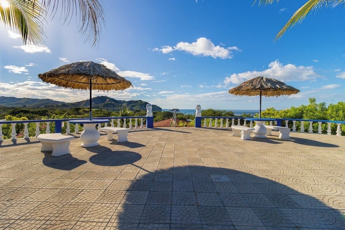 Una terraza exterior con sombrillas de paja, mesas y bancos de piedra blanca, con vistas a un paisaje montañoso.
