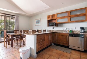 A kitchen with wooden cabinets, a microwave, a dishwasher, and a dining area with chairs and a table. - Resort Pool and Sunset Views: 2BR in Playa del Coco (Playa del Coco)