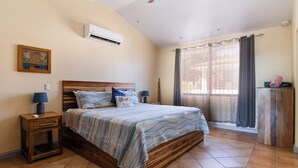 A bedroom with a wooden bed, a nightstand, a lamp, a wall-mounted air conditioner, and a window with curtains. - Resort Pool and Sunset Views: 2BR in Playa del Coco (Playa del Coco)