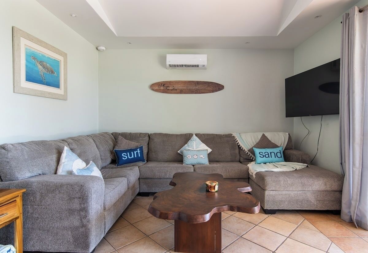 A living room with a sectional sofa, a wooden coffee table, a mounted TV, and an air conditioning unit.