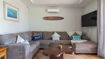A living room with a sectional sofa, a wooden coffee table, a mounted TV, and an air conditioning unit.