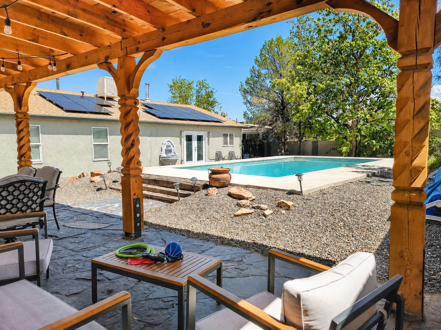 A covered patio area with outdoor seating, a swimming pool, solar panels, and a house in the background.