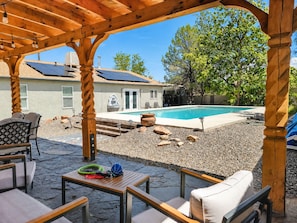 A covered patio area with outdoor seating, a swimming pool, solar panels, and a house in the background.