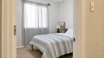 Un dormitorio con cama, mesita de noche, lámpara, ventilador de techo y ventana con cortinas.