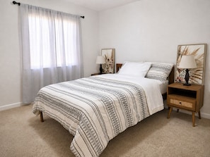 A bedroom with a bed, two nightstands, a window with sheer curtains, and wall art.