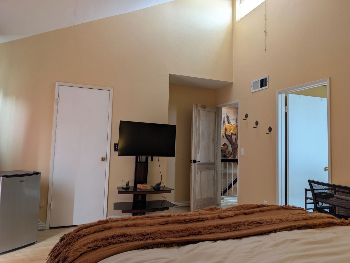 A bedroom with a bed, a television, a small refrigerator, and a door leading to another room.