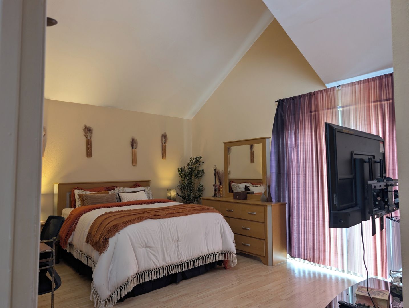 A bedroom with a bed, dresser, mirror, television, and a window with curtains.