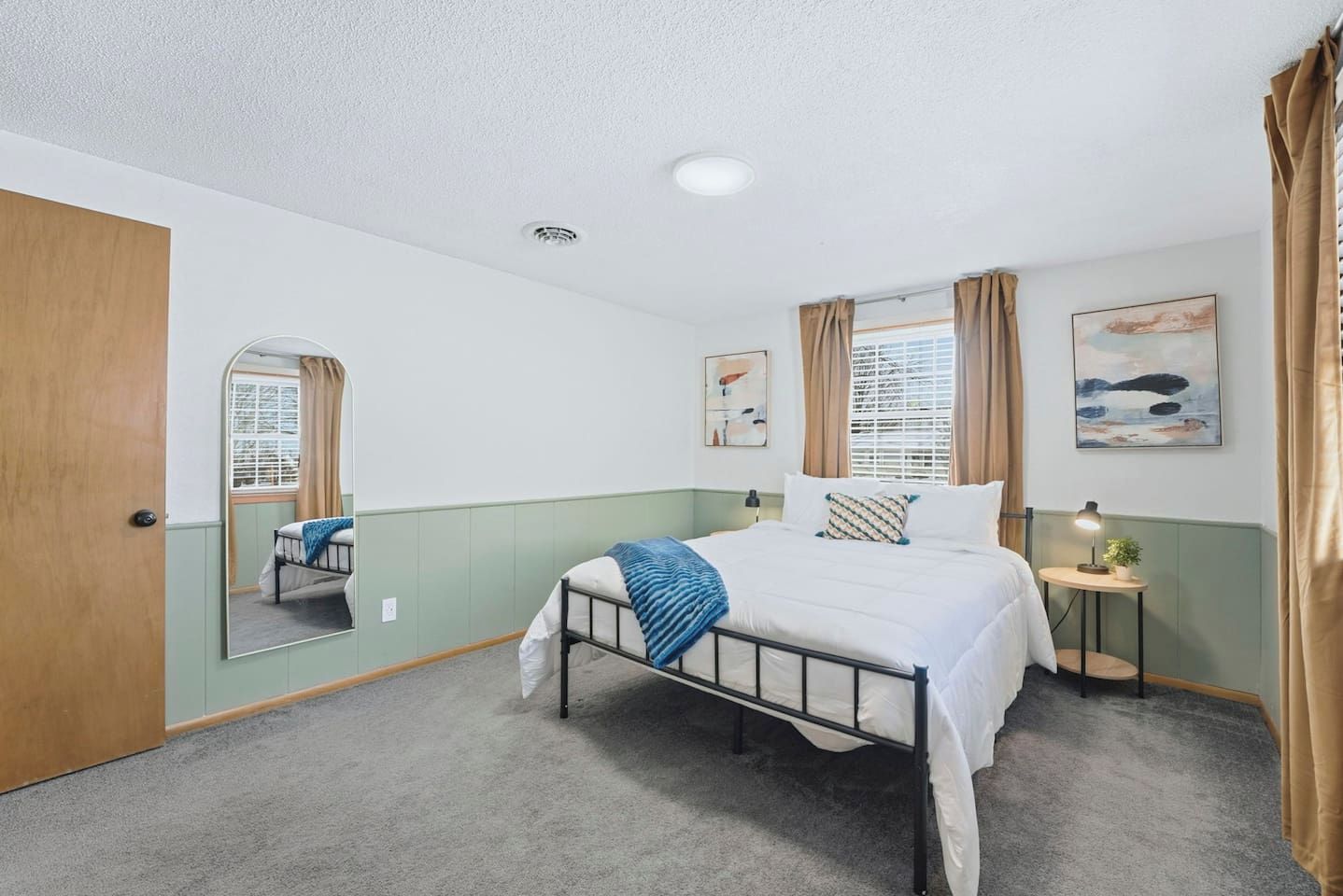 A bedroom with a single bed, a bedside table with a lamp, a window with curtains, and a mirror on the wall.