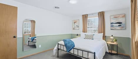 A bedroom with a single bed, a bedside table with a lamp, a window with curtains, and a mirror on the wall.