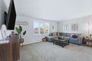 A living room with a grey sofa, a coffee table, a flat-screen TV, and a large window with blinds. - Huntertown Haven |3 Bed 3 Ba Ranch Families & Pets (Huntertown)