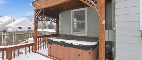 A hot tub on a covered porch with a wooden railing and a window.