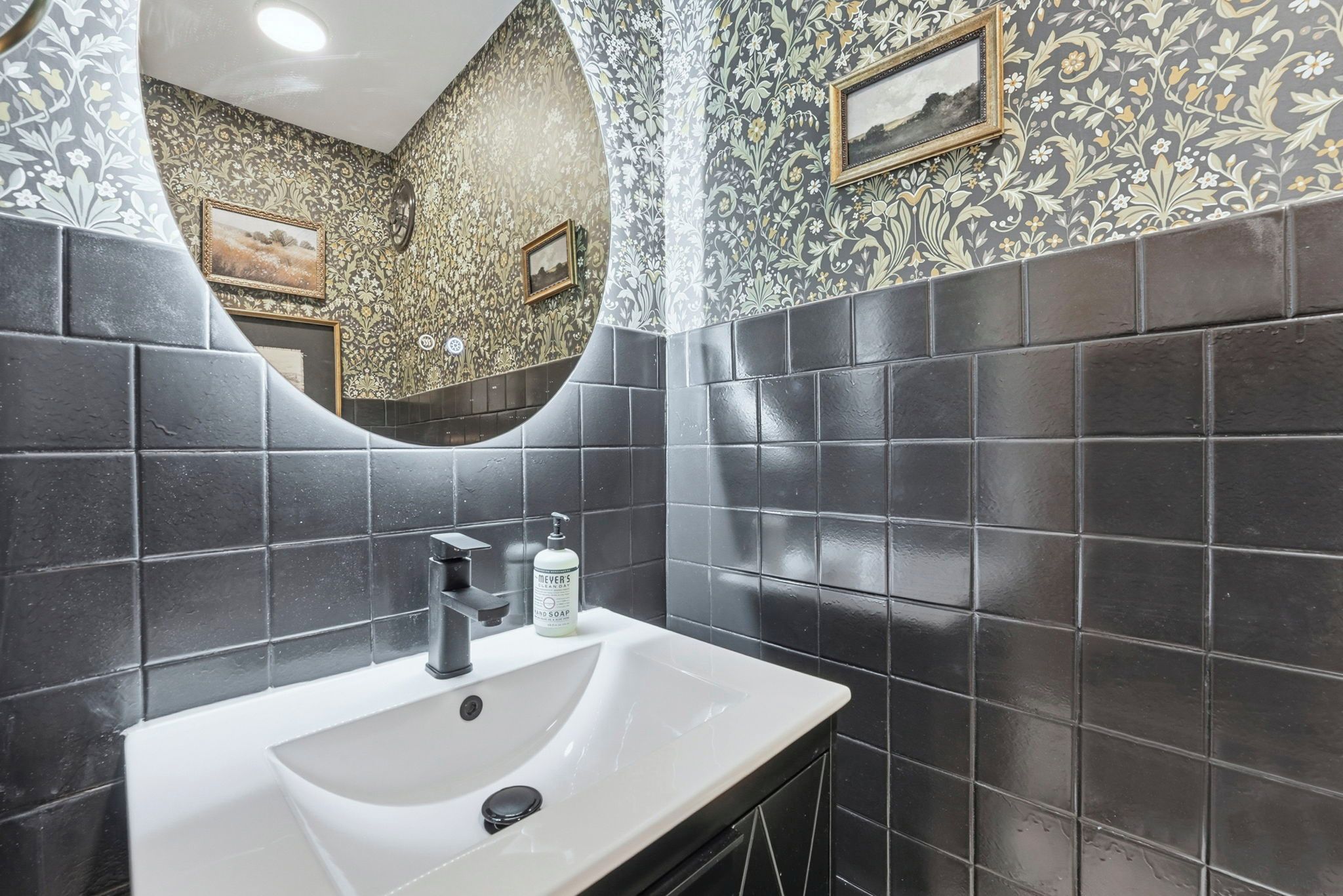 Un baño con lavamanos blanco, baldosas negras, un espejo y una botella de jabón.