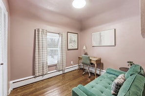 A room with a wooden floor, a green sofa, a desk with a chair, a window with curtains, and a framed picture on the wall. - 2926 Smith Road House (Fort Wayne)