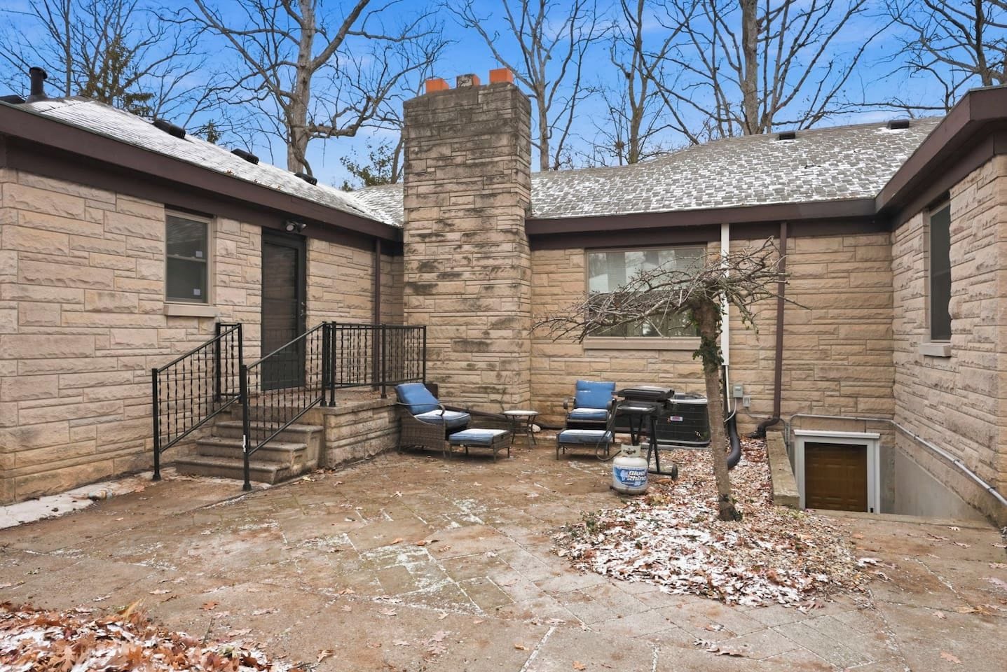 A stone building with a brick chimney, a covered patio with outdoor furniture, and a small tree.