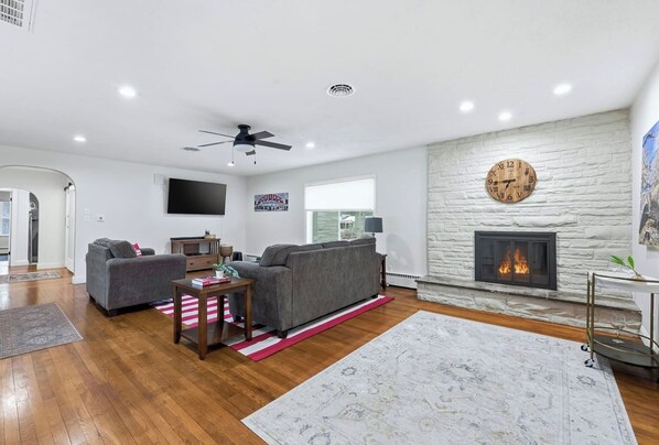 A modern living room with a fireplace, a ceiling fan, and a large clock on the wall. - Hoosier House! Massive 4 BD 2.5Ba for 12 TVs (Fort Wayne)