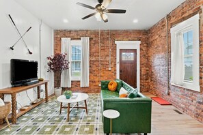 A living room with a green sofa, a television on a wooden stand, a ceiling fan, and a brick wall. - 2BR | Tennis, Kitchen, PetsWelcome, Fenced Yard (Fort Wayne)