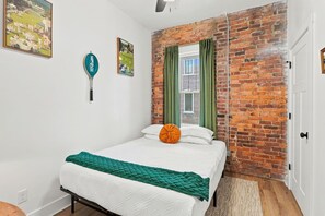 A bedroom with a brick wall, a bed with green and white bedding, a window with green curtains, and a wall-mounted lamp. - 2BR | Tennis, Kitchen, PetsWelcome, Fenced Yard (Fort Wayne)