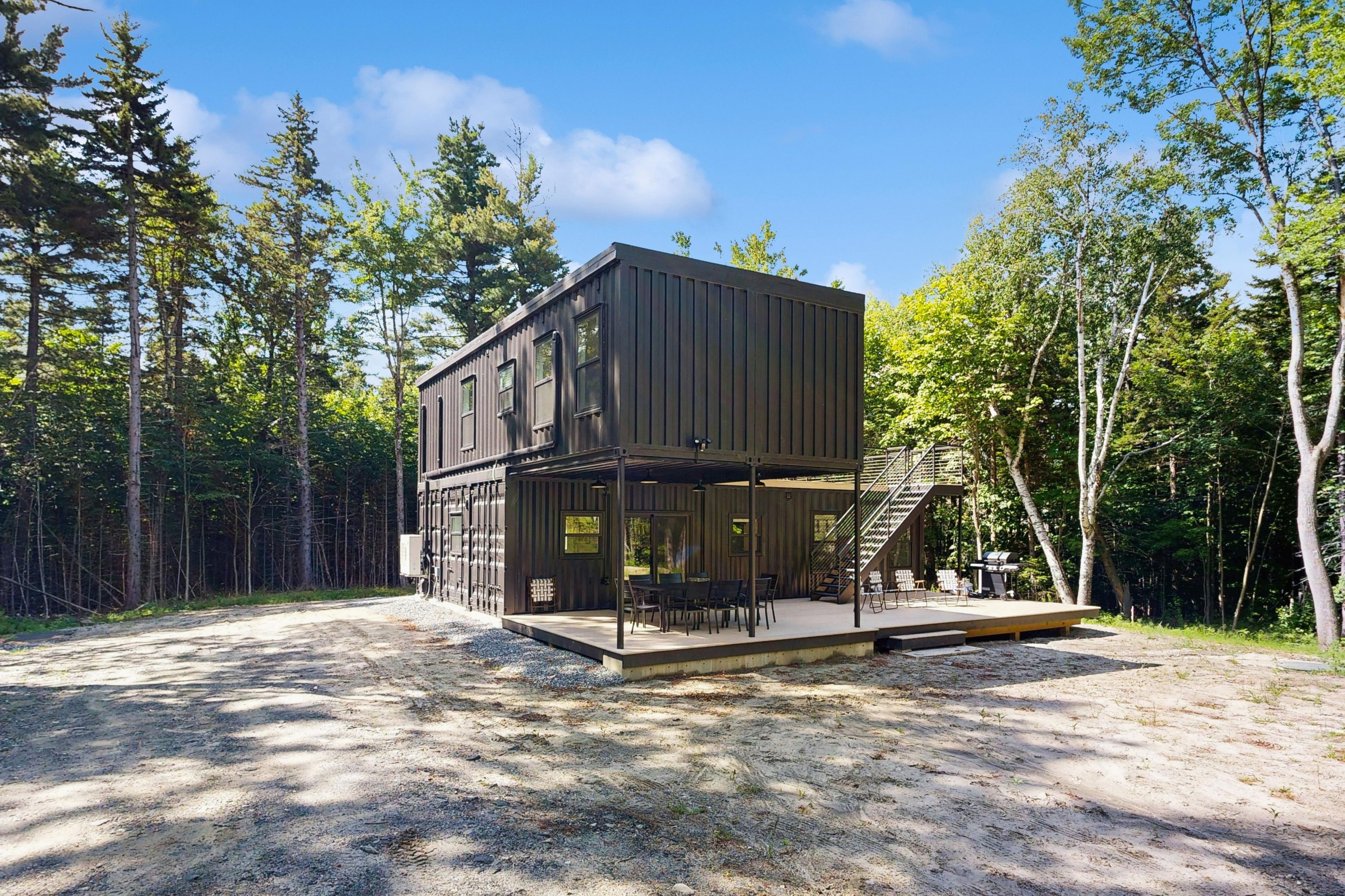 Eine moderne, zweigeschossige Holzhütte mit einer Terrasse und Sitzgelegenheit im Freien, umgeben von einem Wald.