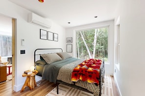Un dormitorio con cama, mesita de noche, ventana con vista a árboles y aire acondicionado instalado en la pared.