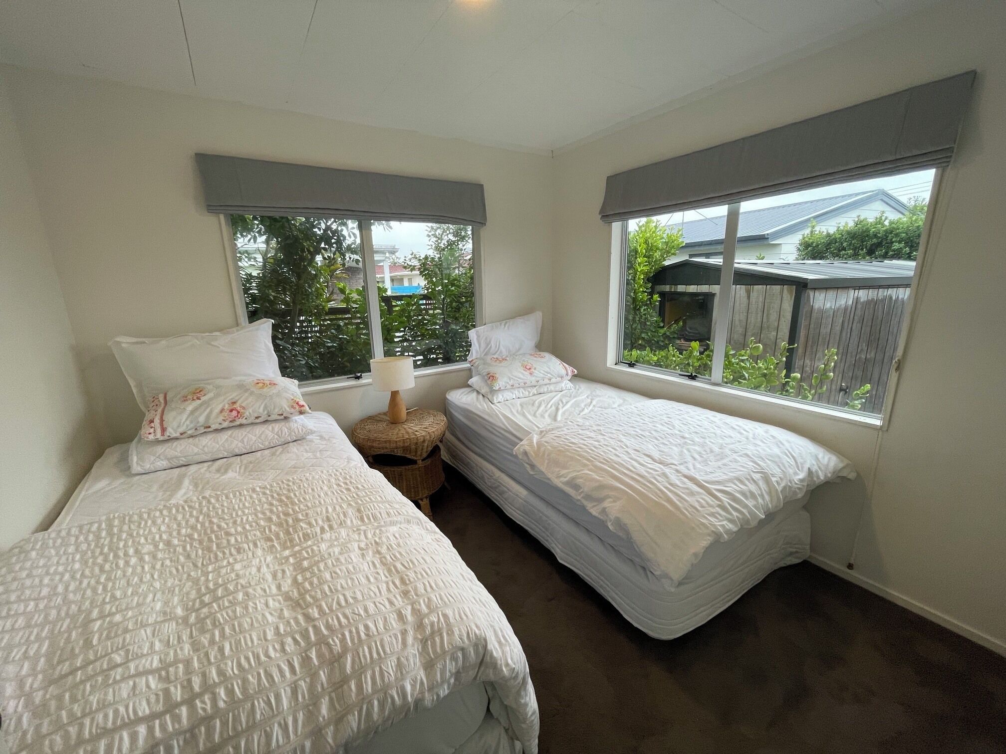 A bedroom with two single beds, a nightstand, a lamp, and large windows with a view of the outside.