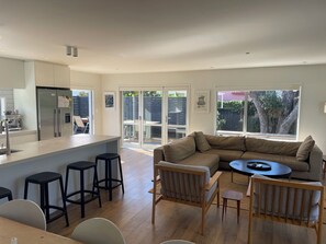 A modern kitchen with a central island, a dining area with a sofa and chairs, and a large window providing a view of the outdoors. - Pye-a-Nui, a peaceful retreat at Whangamata beach. (Whangamatā)