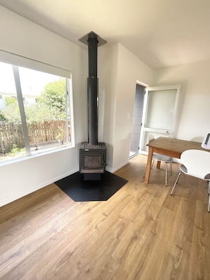 A room with a wood-burning stove, a dining table with chairs, and a window with a view of a fence and trees. - Pye-a-Nui, a peaceful retreat at Whangamata beach. (Whangamatā)