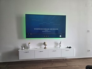 Ein modernes Wohnzimmer mit einem an der Wand befestigten Flachbildfernseher, der ein Musikvideo zeigt. Darunter befindet sich eine weiße Kommode mit Schubladen und Dekoration.