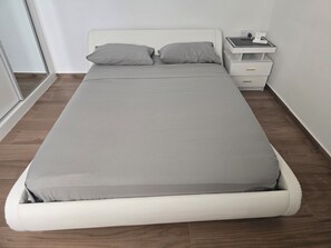 Ein Einzelbett mit grauer Matratze und Kissen, ein Nachttisch mit Telefon und Ladegerät sowie eine Kommode.