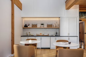 Una cocina moderna con armarios blancos, encimera negra y techo de madera. Hay dos mesas redondas con sillas, y una pequeña isla con un fregadero.