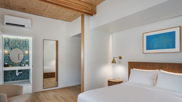 Un dormitorio moderno con una cama grande, cabecera de madera, mesita de noche con lámpara, un espejo amplio y un baño con pared de azulejos y lavamanos.