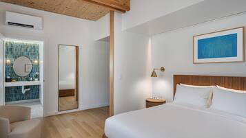 A modern bedroom with a large bed, a wooden headboard, a bedside table with a lamp, a large mirror, and a bathroom with a tiled wall and sink.