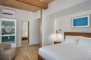 A modern bedroom with a large bed, a wooden headboard, a bedside table with a lamp, a large mirror, and a bathroom with a tiled wall and sink.