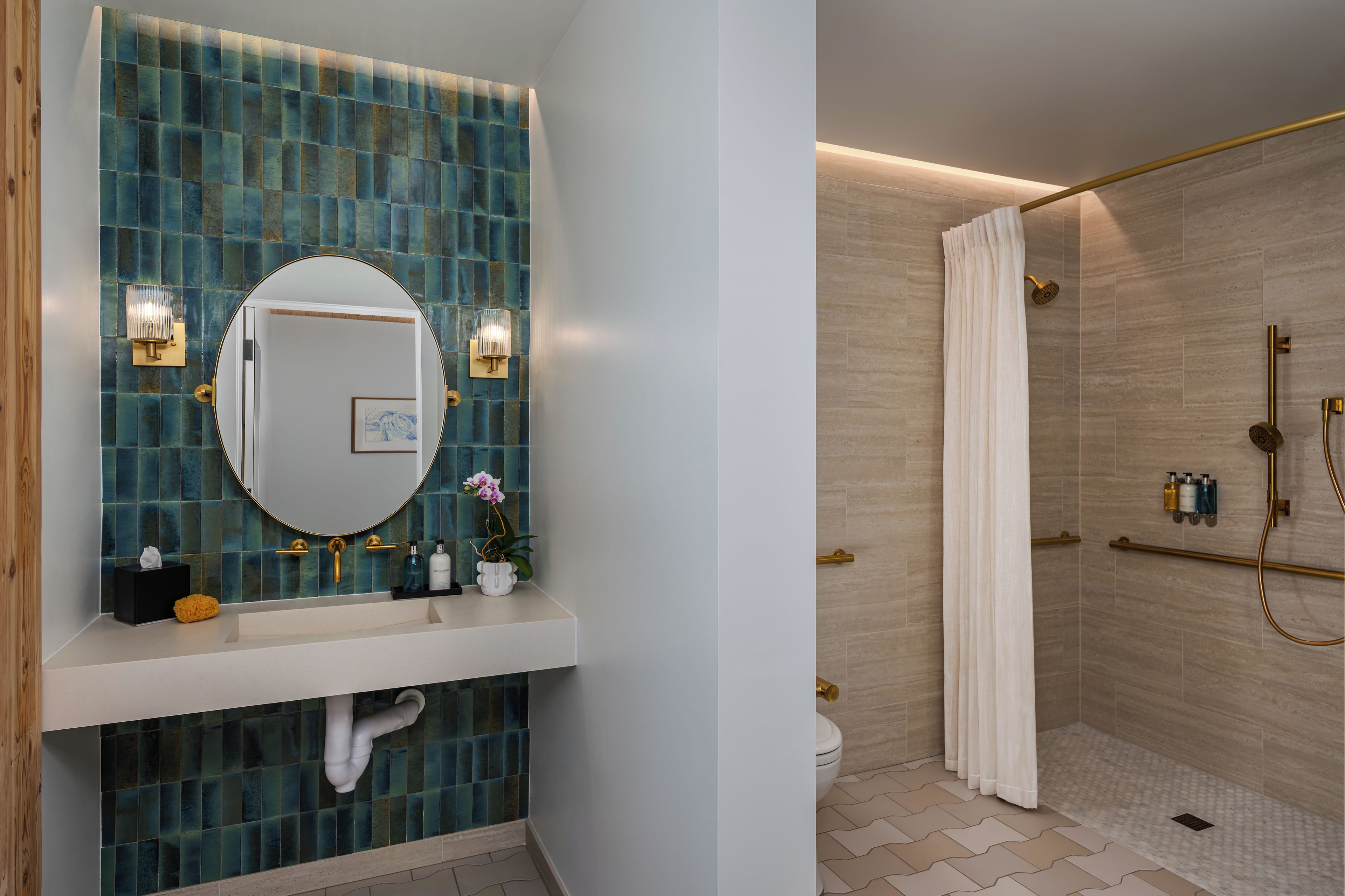 Un baño con pared de azulejos azules, un espejo redondo, dos luces empotradas en la pared, un lavabo blanco, un inodoro y una ducha con cabezal dorado y cortina.