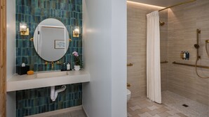 Un baño con pared de azulejos azules, un espejo redondo, dos luces empotradas en la pared, un lavabo blanco, un inodoro y una ducha con cabezal dorado y cortina.