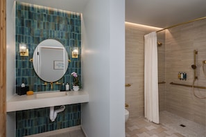 Un baño con pared de azulejos azules, un espejo redondo, dos luces empotradas en la pared, un lavabo blanco, un inodoro y una ducha con cabezal dorado y cortina.