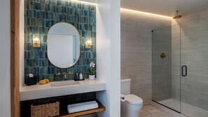 Un baño con pared de azulejos azules, un lavamanos blanco, un espejo redondo, inodoro, cabina de ducha con mampara de cristal y un estante de madera con toallas y artículos de aseo.