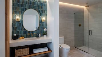A bathroom with a blue-tiled wall, a white sink, a round mirror, a toilet, a glass-enclosed shower, and a wooden shelf with towels and toiletries.