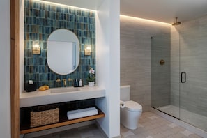 A bathroom with a blue-tiled wall, a white sink, a round mirror, a toilet, a glass-enclosed shower, and a wooden shelf with towels and toiletries.