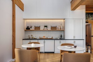 A modern kitchen with white cabinetry, a black countertop, and a wooden ceiling. There are two round tables with chairs, and a small island with a sink.