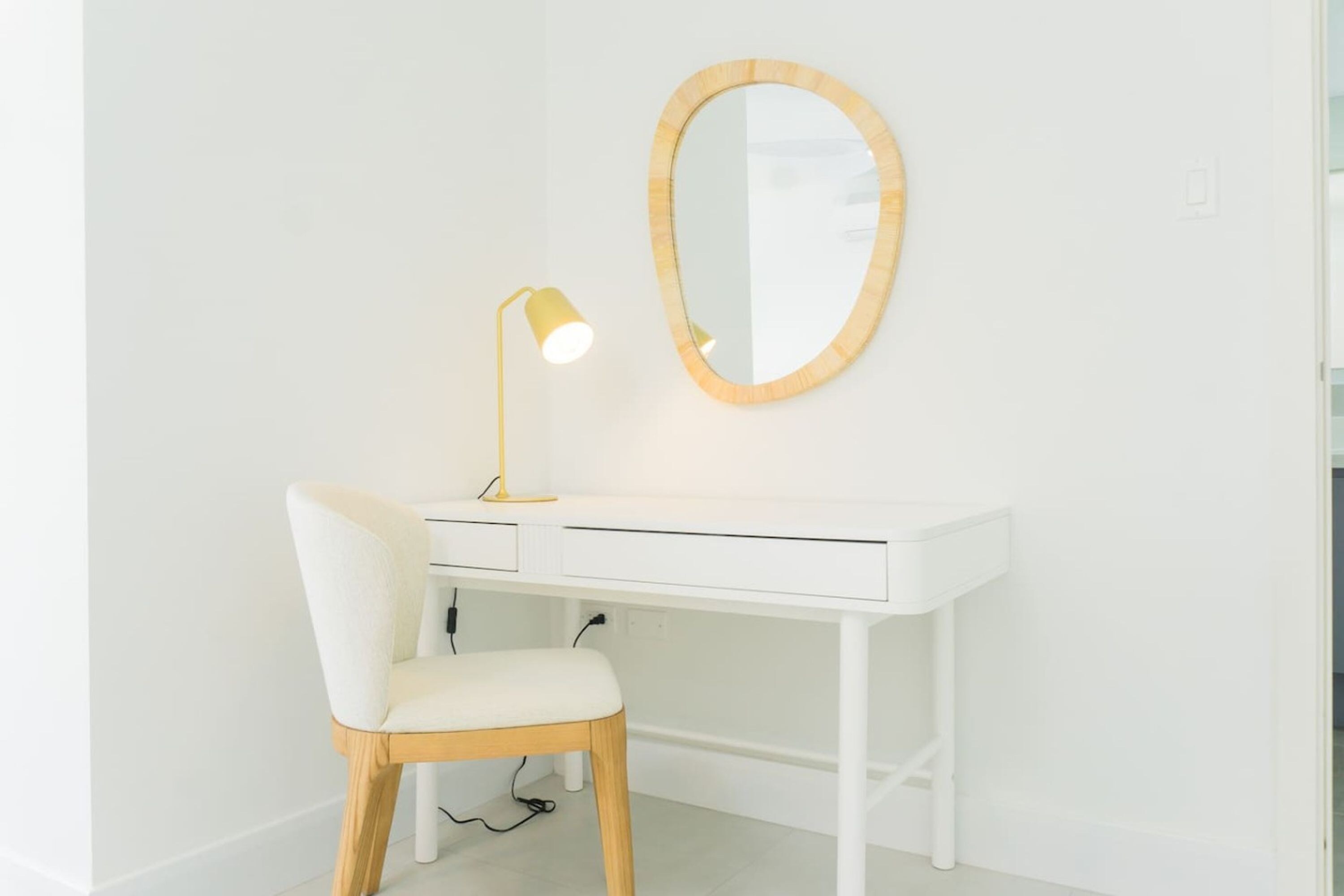 Un bureau blanc avec un miroir et une lampe, une chaise blanche et un câble relié au bureau.