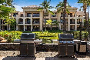 A patio area with two Genesis portable grills, a small pond, and a multi-story building with balconies in the background. - 🏖️Ground-Floor Waikoloa Condo | Corner Unit + Spacious Lanai (Waikoloa)